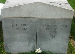 Shared headstone of Joe Mitchell (1849 to 1912) and Aurelia Mitchell (1859 to 1940) at Our Mother of Mercy Catholic Cemetery in Ames, Liberty County, Texas. The double-gabled granite stone shows their names and dates engraved side by side with a single white flower resting at the base.