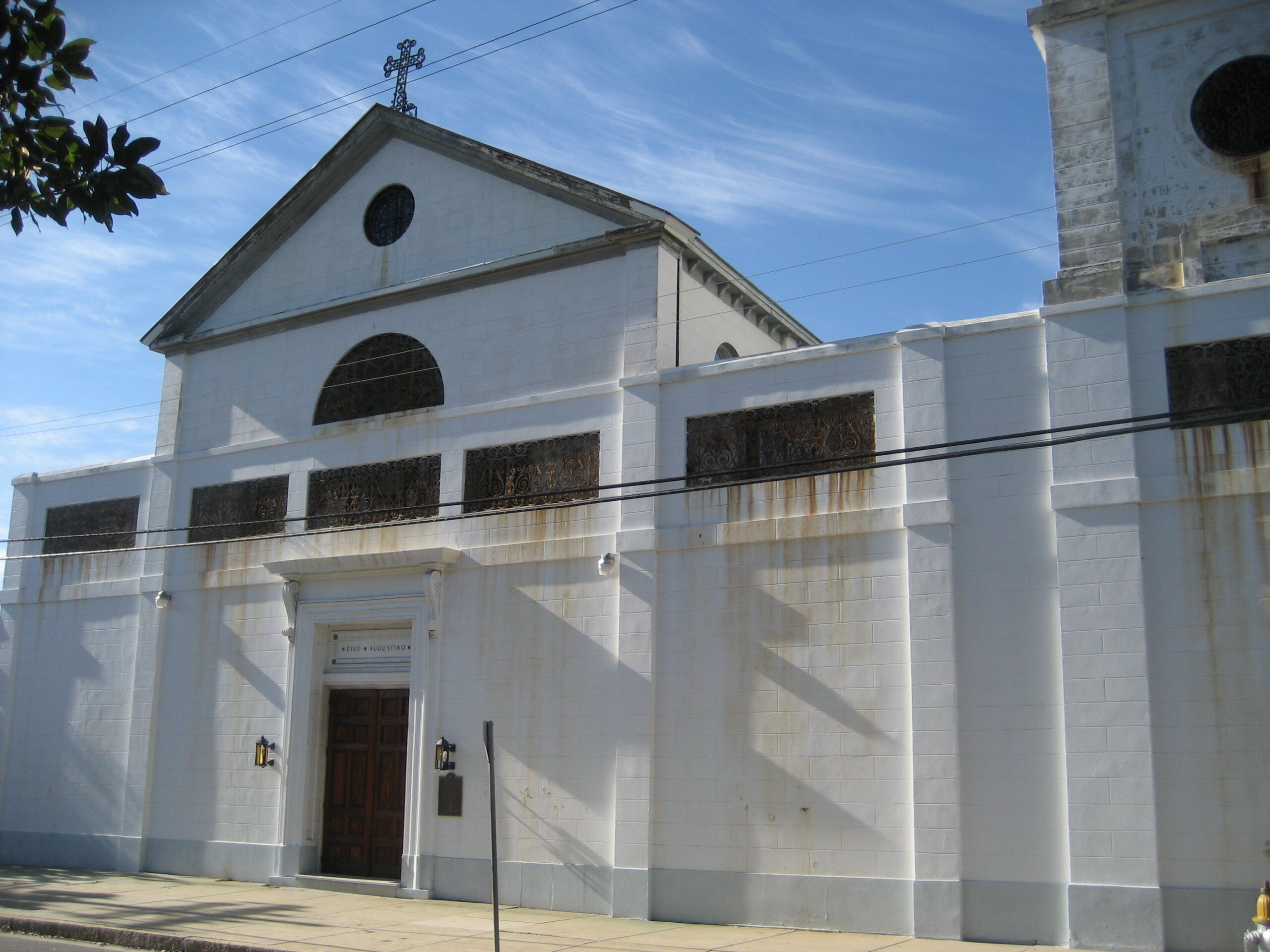 The white stucco front facade of St. Augustine Church in Tremé, New Orleans, with a rooftop cross and a steeple at right against a blue sky.